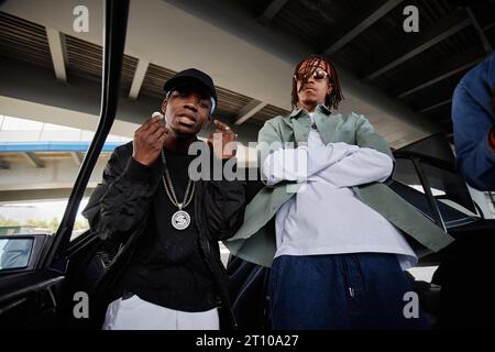 Jeune homme afro-américain en casquette et blouson noir et son copain en lunettes de soleil et bandeau debout près de la voiture devant la caméra Banque D'Images