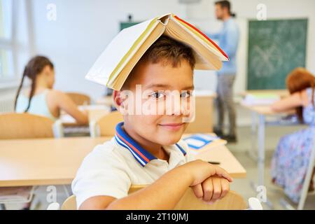 Portrait d'écolier souriant équilibrant le livre sur la tête tout en étant assis dans la salle de classe Banque D'Images