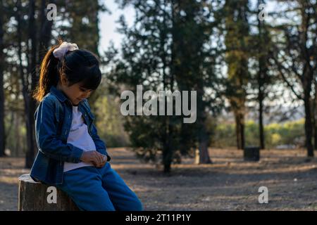 Petite fille latine regardant vers le bas appuyée sur une bûche dans la forêt. Banque D'Images