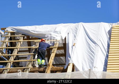 Charpentier remplace les structures de toit endommagées de la vieille maison en bois, filet de sécurité blanc, fond de ciel bleu Banque D'Images