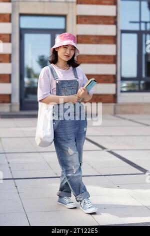 Portrait vertical pleine longueur d'une jeune femme asiatique amusante portant une combinaison en denim comme étudiante sur le campus de l'université souriant à la caméra Banque D'Images