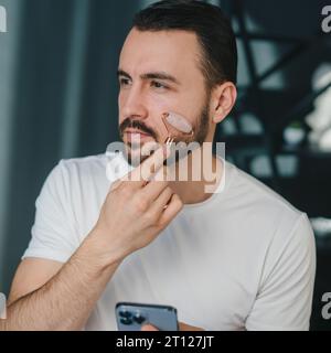 Homme utilisant un rouleau de jade dans sa routine de soins de la peau, massant son visage pour une peau éclatante. Beau jeune homme pratiquant dans l'auto-soin anti-âge avec un Banque D'Images