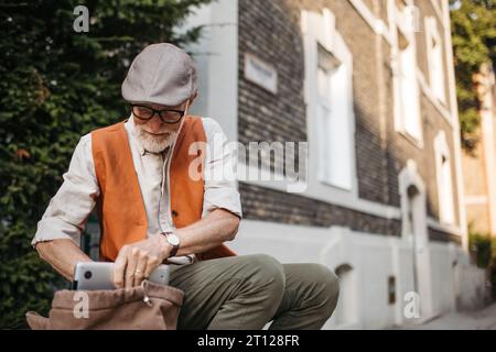 Homme senior assis sur le trottoir de la rue dans la ville. Portrait d'un homme âgé cherchant des choses dans un sac à dos. Banque D'Images