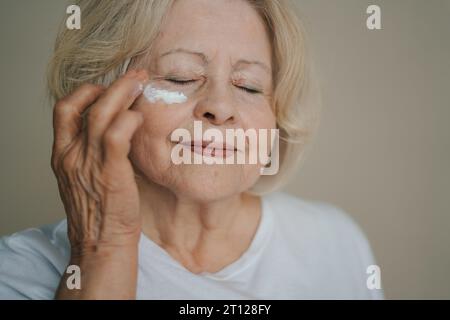 Belle femme senior appliquant la crème pour le visage sous ses yeux sur fond de studio. Dame âgée prenant soin de sa peau. Concept de beauté mature. Banque D'Images