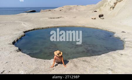 Femme seule en maillot de bain portant un chapeau assis sur le bord d'un petit étang près de la plage à Cane Malu, en Sardaigne, Italie. Vacances de voyage Banque D'Images