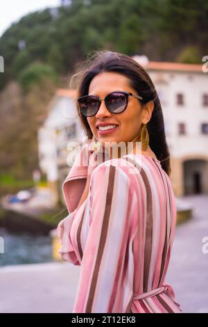 Portrait d'une jeune femme latine dans une robe rose et des lunettes de soleil profitant des vacances d'été dans une belle ville côtière, posant au bord de la mer, à la verticale Banque D'Images