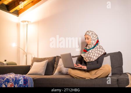 Fille arabe avec un voile blanc à son ordinateur à la maison sur le canapé dans son salon, faisant un travail. Jeune homme arabe moderne avec les nouvelles technologies Banque D'Images