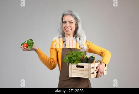 Heureuse femme âgée fière en tablier tenir une boîte en bois avec des légumes écologiques, profitez de la récolte Banque D'Images
