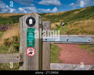 Signalisation bilingue sur un portail en bois en gallois et en anglais donnant des informations aux marcheurs sur le sentier côtier du Pembrokeshire au pays de Galles, Royaume-Uni. Un seul marcheur A. Banque D'Images