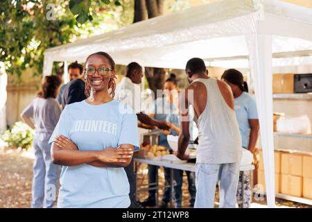 Jeune femme noire avec des lunettes debout à l'extérieur, les bras croisés, regardant la caméra. Un groupe diversifié de bénévoles soutient un programme à but non lucratif dédié au soulagement de la faim et à l'aide aux personnes nécessiteuses. Banque D'Images