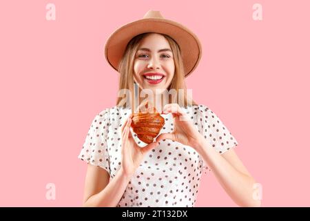 Belle jeune femme dans le chapeau en osier avec croissant savoureux sur fond rose Banque D'Images