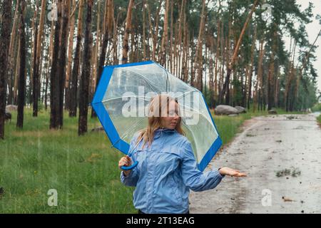 Fille blonde vérifiant le temps sous un parapluie transparent à l'extérieur dans le parc. Heure d'automne pluvieux mauvaises conditions de prévision saison. Touriste avec parapluie voyage dans de nouveaux endroits sous la pluie. Paysage Banque D'Images