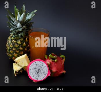 Un grand verre de jus de fruits fraîchement pressés sur fond noir, à côté d'un ananas mûr et d'un fruit pitaya coupé. Gros plan. Banque D'Images