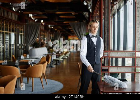 Portrait d'un jeune homme élégant portant un nettoyage uniforme de smoking avec plumeau à l'intérieur d'un restaurant de luxe, espace de copie Banque D'Images