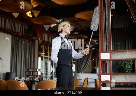 Portrait de vue latérale de jeune homme élégant comme serveur portant smoking uniforme nettoyage avec plumeau dans le restaurant de luxe, copie espace Banque D'Images