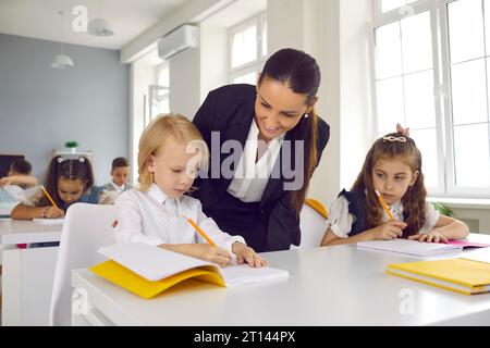 Enseignante amicale observe et aide le petit garçon avec son travail scolaire pendant la leçon. Banque D'Images