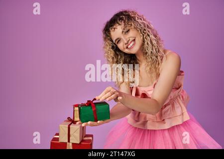 joyeuse femme blonde en costume rose tenant l'un des cadeaux et souriant joyeusement à la caméra Banque D'Images