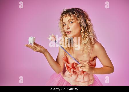 belle femme blonde en costume de fée de dent jouant sort sur la dent de bébé avec sa baguette magique Banque D'Images