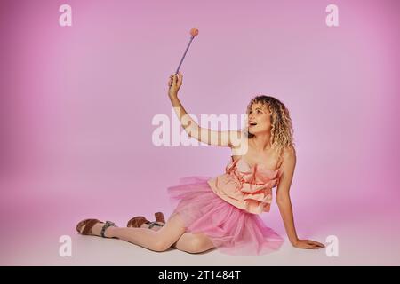 blonde joyeuse avec les cheveux bouclés posant sur fond rose en costume de fée de dent avec baguette magique Banque D'Images