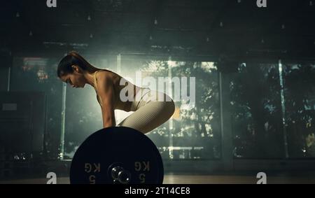 Jeune femme faisant le soulevé de terre avec la barre lourde dans la salle de gym, athlète féminine forte avec le corps musclé soulevant des poids, l'exercice avec barre. Banque D'Images