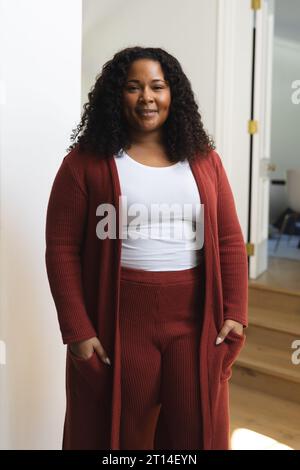 Heureuse femme afro-américaine avec les cheveux bouclés debout et souriant dans la maison ensoleillée Banque D'Images