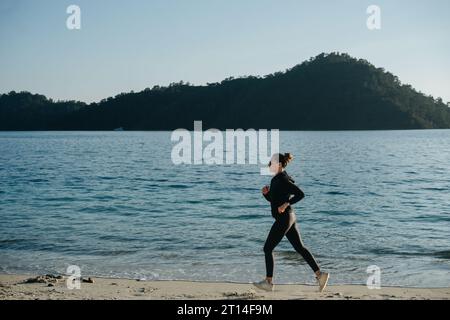 Une femme court le long du sable en sportswear noir sur fond de mer Banque D'Images