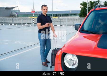 Un homme avec des bagages et un téléphone à la main près de la voiture dans un parking ouvert. Affaires, voyage d'affaires, concept de voyage Banque D'Images