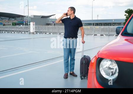 Un jeune homme avec une valise à la main parle au téléphone dans le parking près de la voiture. Concept de voyage Banque D'Images