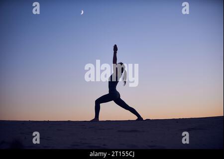 Belle jeune femme pratique yoga asana Virabhadrasana 1 - guerrier pose 1 dans le désert la nuit. Banque D'Images