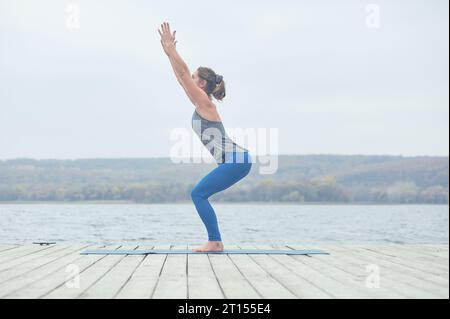 Belle jeune femme pratique yoga asana Utkatasana - chaise pose sur la terrasse en bois près du lac. Banque D'Images