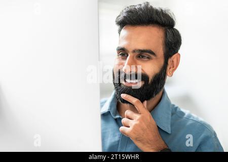 Heureux homme d'affaires indien travaillant sur l'ordinateur dans le bureau, regardant le moniteur et souriant, touchant le menton, gros plan Banque D'Images