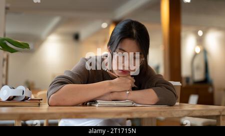 Une jeune femme asiatique fatiguée et pensive aux lunettes se sent ennuyée en lisant un livre ou en faisant ses devoirs dans un café. Banque D'Images