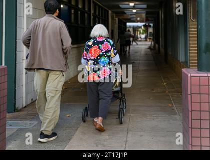 Femme âgée marchant à l'aide d'une marchette à mobilité réduite dans une ruelle commerçante, accompagnée de son fils. Californie. Banque D'Images