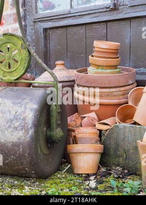 Gros plan sur de vieux outils de jardin, y compris un rouleau à gazon en fonte et des pots en terre cuite vintage empilés par un hangar dans un jardin britannique Banque D'Images