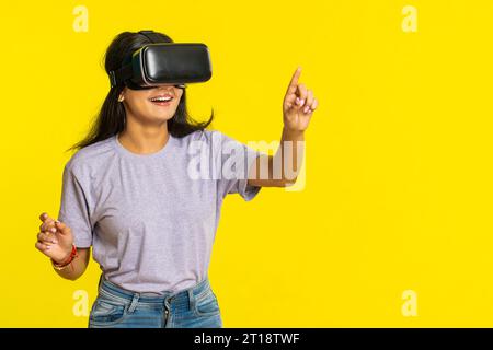 Excité heureuse jeune femme indienne utilisant l'application casque casque casque pour jouer au jeu de simulation. Regarder une vidéo 3D 360 en réalité virtuelle. Fille arabe dans les lunettes VR isolé sur fond jaune. Technologie du futur Banque D'Images