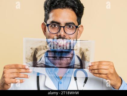 Jeune médecin indien orthodontiste examine une image panoramique de radiographie des dents de la mâchoire. Modèle 3D de la bouche du patient, IRM. Dentisterie, soins bucco-dentaires. Homme stomatologique arabe sur fond beige Banque D'Images