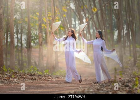 Deux femmes en robes tuniques vietnamiennes traditionnelles (ao dai) et chapeaux (non la) marchant dans une forêt, Vietnam Banque D'Images