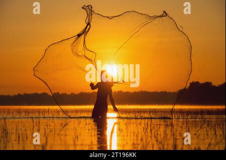 Silhouette d'un pêcheur debout dans une rivière jetant un filet de pêche au coucher du soleil, Thaïlande Banque D'Images