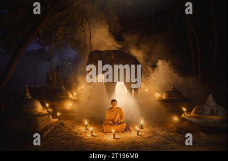 Moine dans une robe orange traditionnelle (kasay) entouré de bougies assis devant un éléphant, Surin, Thaïlande Banque D'Images