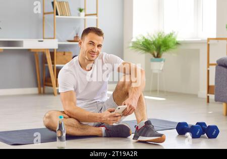Heureux, homme en forme se détendant après l'entraînement à la maison, tenant le téléphone portable et regardant la caméra Banque D'Images