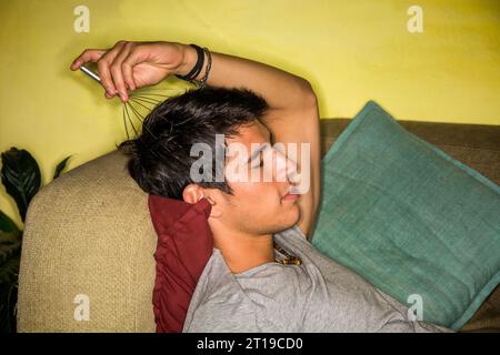 Un homme allongé sur un canapé avec un masseur de tête dans sa main.photo d'un jeune et bel homme se détendant sur un canapé avec un masseur de tête en main Banque D'Images