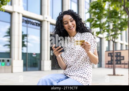 Jeune belle femme marchant dans la ville, femme latino-américaine satisfaite avec les cheveux bouclés tient le téléphone et la carte de crédit bancaire dans les mains, fait des achats en ligne heureusement livres services et choisit en ligne. Banque D'Images