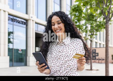 Jeune belle femme marchant dans la ville, femme latino-américaine satisfaite avec les cheveux bouclés tient le téléphone et la carte de crédit bancaire dans les mains, fait des achats en ligne heureusement livres services et choisit en ligne. Banque D'Images