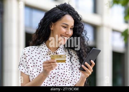 Jeune belle femme marchant dans la ville, femme latino-américaine satisfaite avec les cheveux bouclés tient le téléphone et la carte de crédit bancaire dans les mains, fait des achats en ligne heureusement livres services et choisit en ligne. Banque D'Images