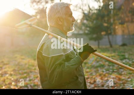 Travailleur de maintenance senior regardant loin, tout en gardant le balai sur l'épaule dans la chaude journée d'automne. Vue latérale de l'homme barbu en âge marchant avec balai, avec fond ensoleillé. Travaux d'entretien, concept d'automne. Banque D'Images