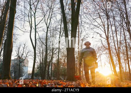 Homme âgé souriant travaillant avec le souffleur lourd de feuilles, laisse briller dans la lumière du soleil agréable. Vue à faible angle d'un homme caucasien barbu en vêtements de travail utilisant un équipement portatif pour enlever les feuilles. Concept de travail. Banque D'Images