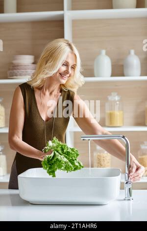 positive femme végétarienne d'âge moyen avec les cheveux blonds lavant la laitue fraîche, tir vertical Banque D'Images