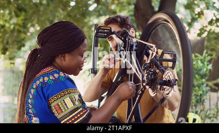 Jeune couple motivé à l'extérieur réparant des composants endommagés du vélo avec un équipement professionnel. Petit ami actif et petite amie réparer le cadre de vélo cassé et le cogset pour le cyclisme de loisirs d'été. Banque D'Images