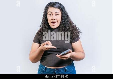 Fille étonné utilisant et pointant le téléphone cellulaire regardant la caméra isolée. Portrait d'une femme latine surprise tenant un téléphone portable et regardant la caméra Banque D'Images