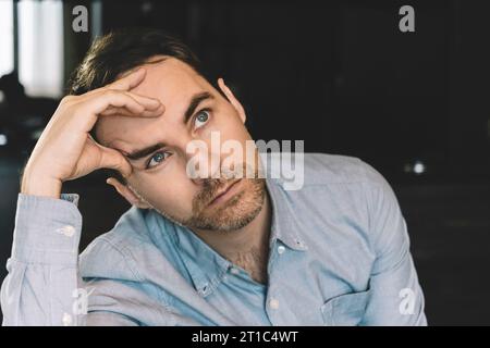 un jeune homme est assis pensif et regarde loin en tenant sa tête avec sa main. Concept de pensées et de rêves Banque D'Images